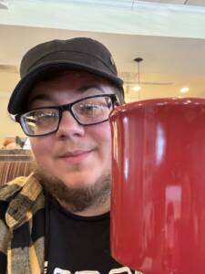 Man wearing glasses and a billed hat, holding a red coffee mug